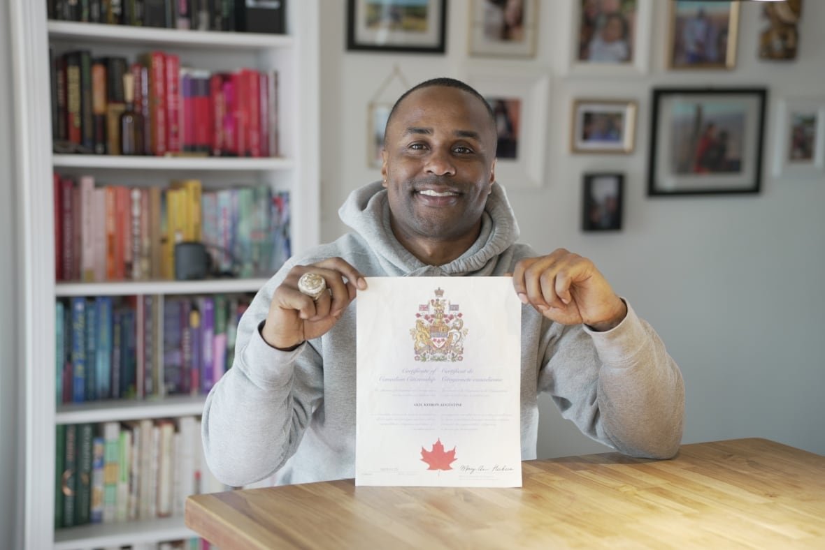 A smiling man wearing a hoodie and a large NBA championship ring on his right hand sits at a wooden table in front of a bookshelf holding a document with the Canadian coat of arms at the top and a large maple leaf at the bottom. 