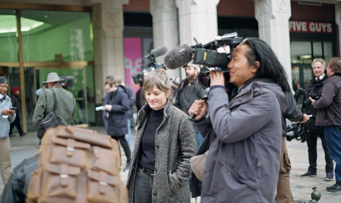 A man carries a film camera on his shoulder and is pointing it at something off screen. A woman smiles beside him. They are outside in some sort of plaza or street and people mill around them. 