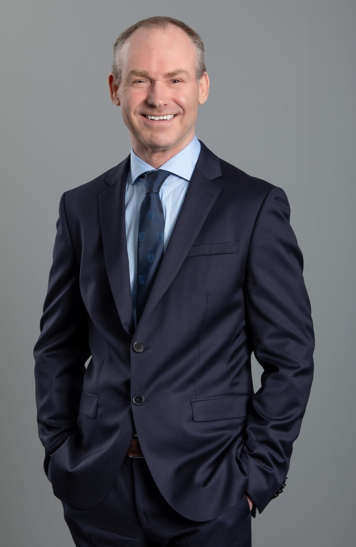 A balding man stands, smiling, wearing a navy suit, tie and shirt. 