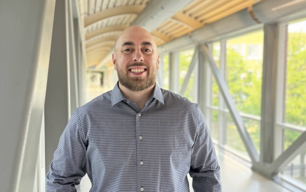 A man in a button-down dress shirt smiles as he stands in a bright indoor walkway with large windows looking over a treed area.