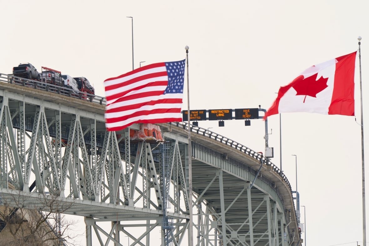 The Canada and U.S. flags fly in the foreground as vehicles travel on a bridge in the background.