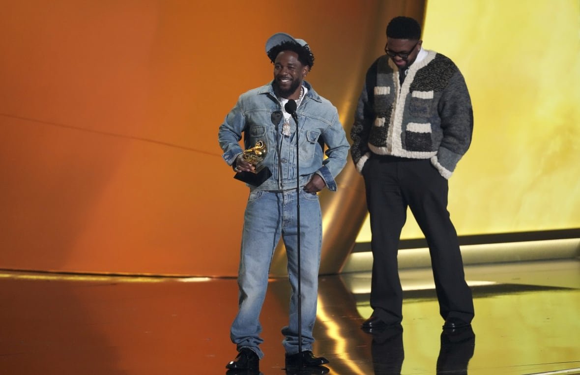 A smiling man wearing all denim stands at a microphone, while another man stands off to the side. 