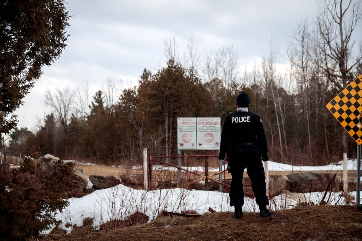 Seen from behind, a police officer looks across a snowy, partly forested landscape. A sign in the middle distance indicates a border crossing.