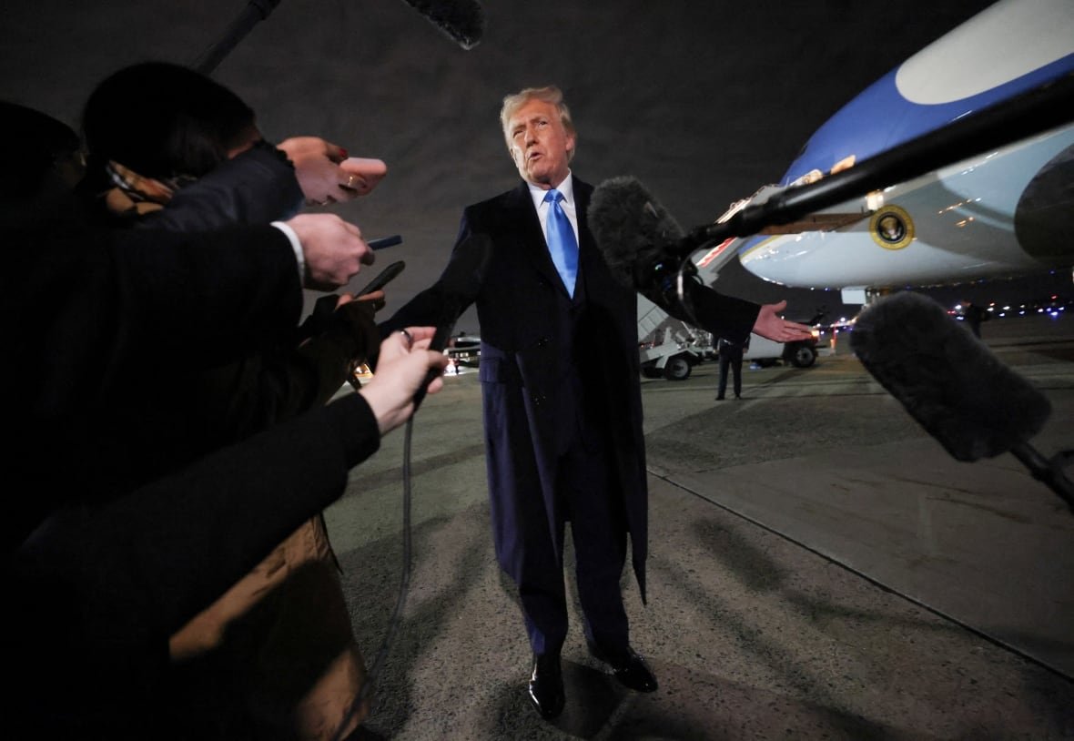 A man speaks on a tarmarc surrounded by reporters.
