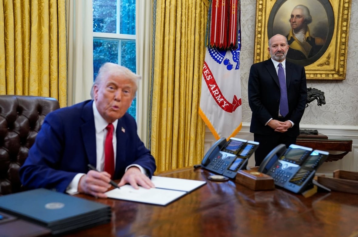 Two men are seen indoors, one sitting and one standing.