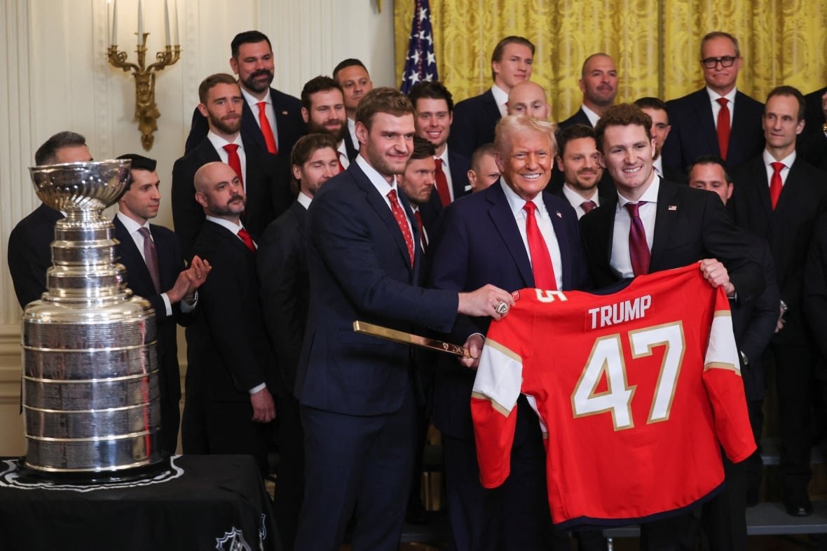 A group of men smile while holding a red jersey that says Trump, with the Stanley Cup to the left.