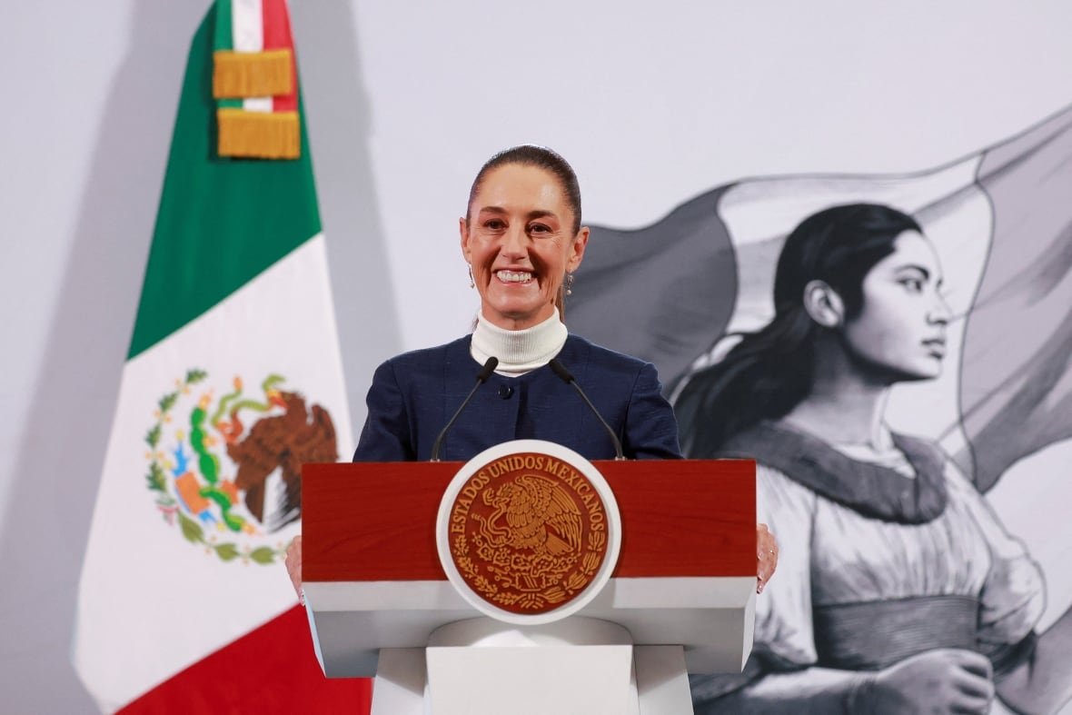 A woman smiles at a podium with the Mexican flag behind her.