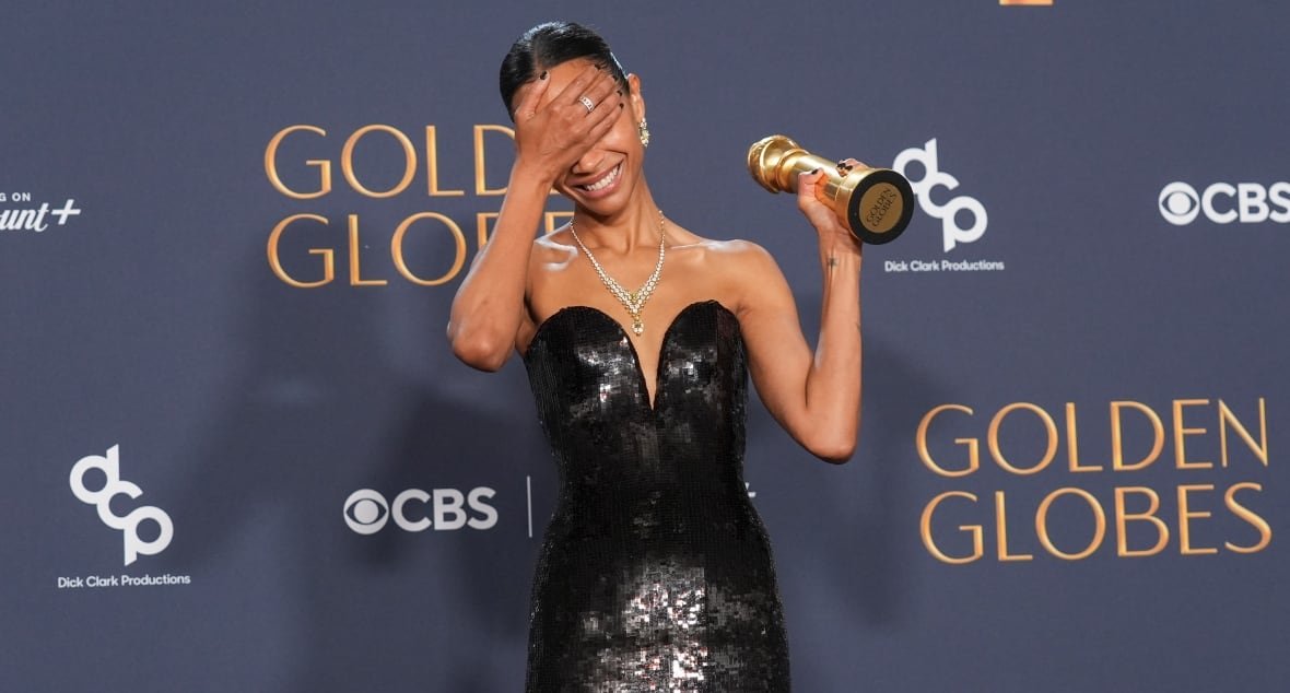 A woman in a sparkly black dress holds up an award in one hand and claps the other hand over her eyes. She is smiling and standing in front of a backdrop that says "Golden Globes" on it.
