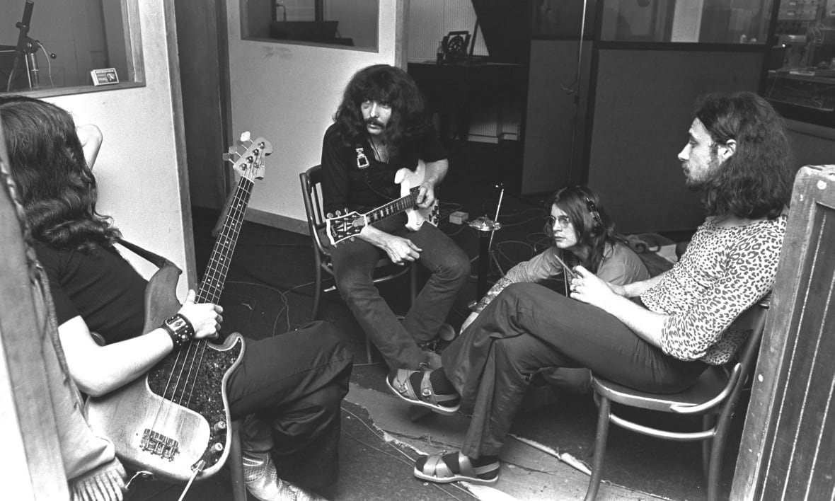 A black and white photo shows three men sitting in a circle on chairs, with a fourth man sitting on the floor. One man is holding a guitar and another is holding a bass.