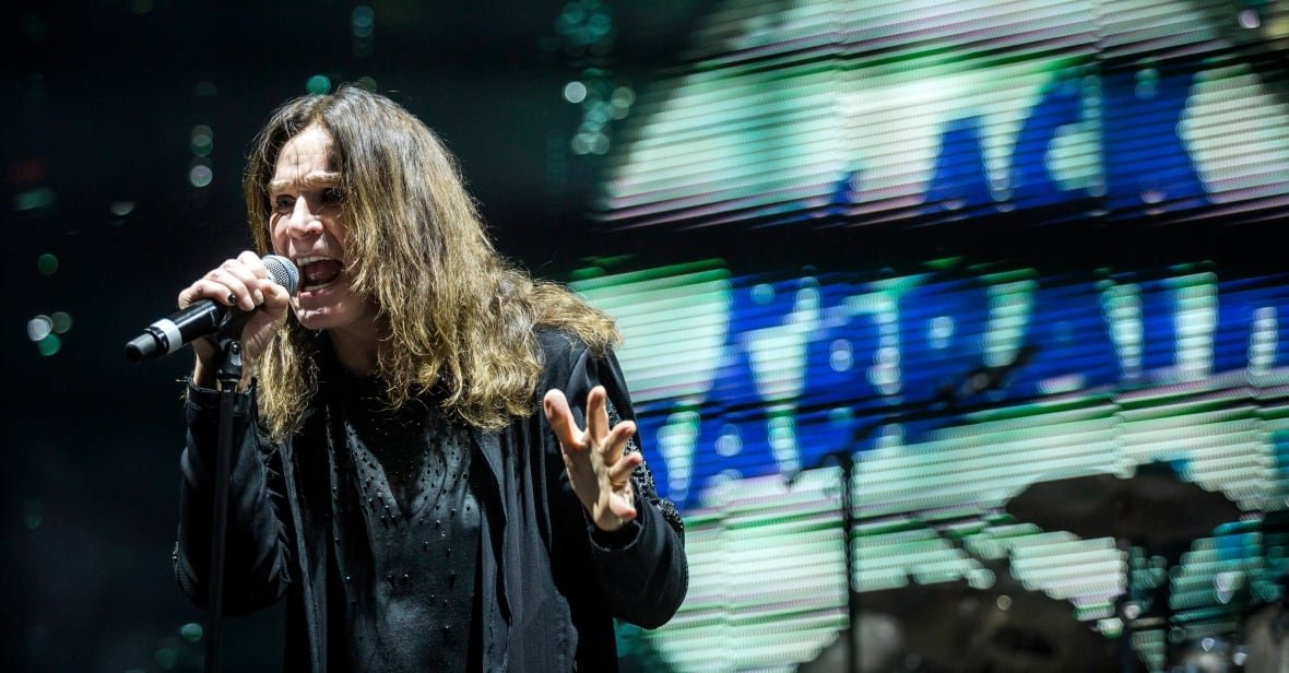 A man with long hair sings into a microphone. Behind him a screen that says Black Sabbath in distorted text can be seen.