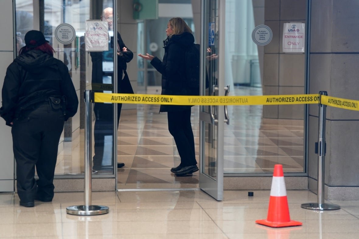 Yellow tape is shown outside a building entrance, with a woman visible inside speaking as a man looks on.