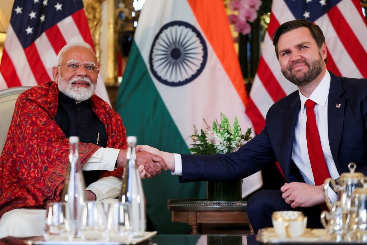 Two men pose for a photo while shaking hands and seated.