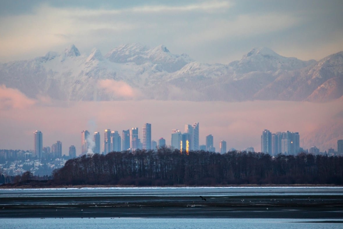A picturesque series of mountain peaks looms over a city skyline, with pink and purple light illuminating the background and trees and a water body in the foreground.