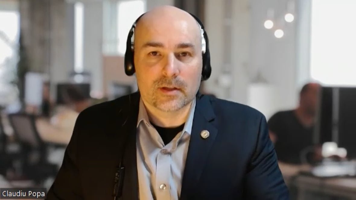 A man wearing a headset and dark suit is shown looking into the camera with an office in the background. 