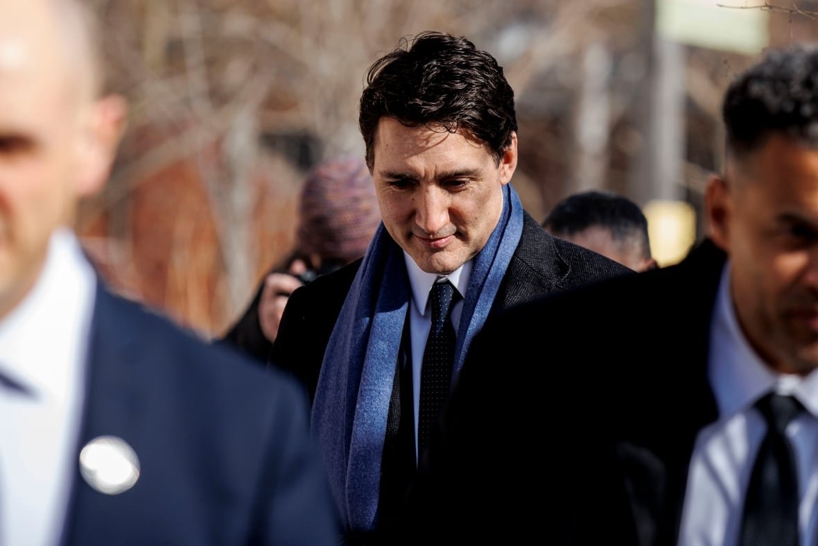 A man with brown hair, a black boat and a blue scarf walks with his head down on a sunny winter day.