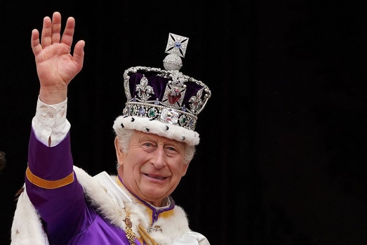 A person wearing a crown holds up their right arm to wave in front of a black background.
