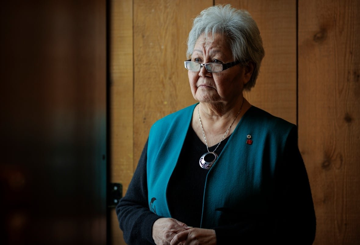 An elderly woman stands for a portrait in front of a wood wall.