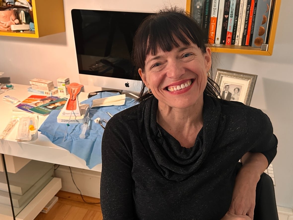 A woman with dark hair and bangs sits in front of a model uterus and medical tools on a table. 