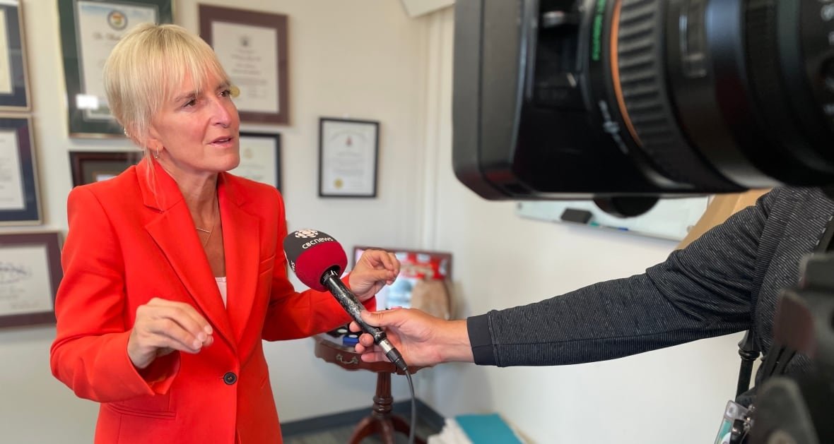 A woman with blonde hair wearing a red suit speaks into a microphone and camera in an office.
