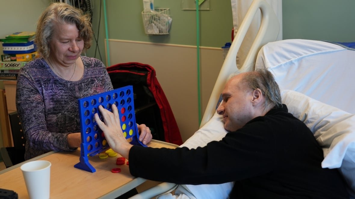 A woman and man play a game on a bedside table in a hospital room. The man is reaching from his hospital bed.
