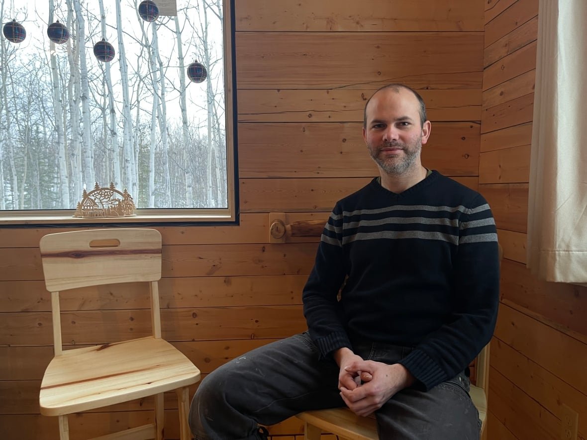 A man sits in a cabin beside a handmade chair. Outside the window is a aspen forest.