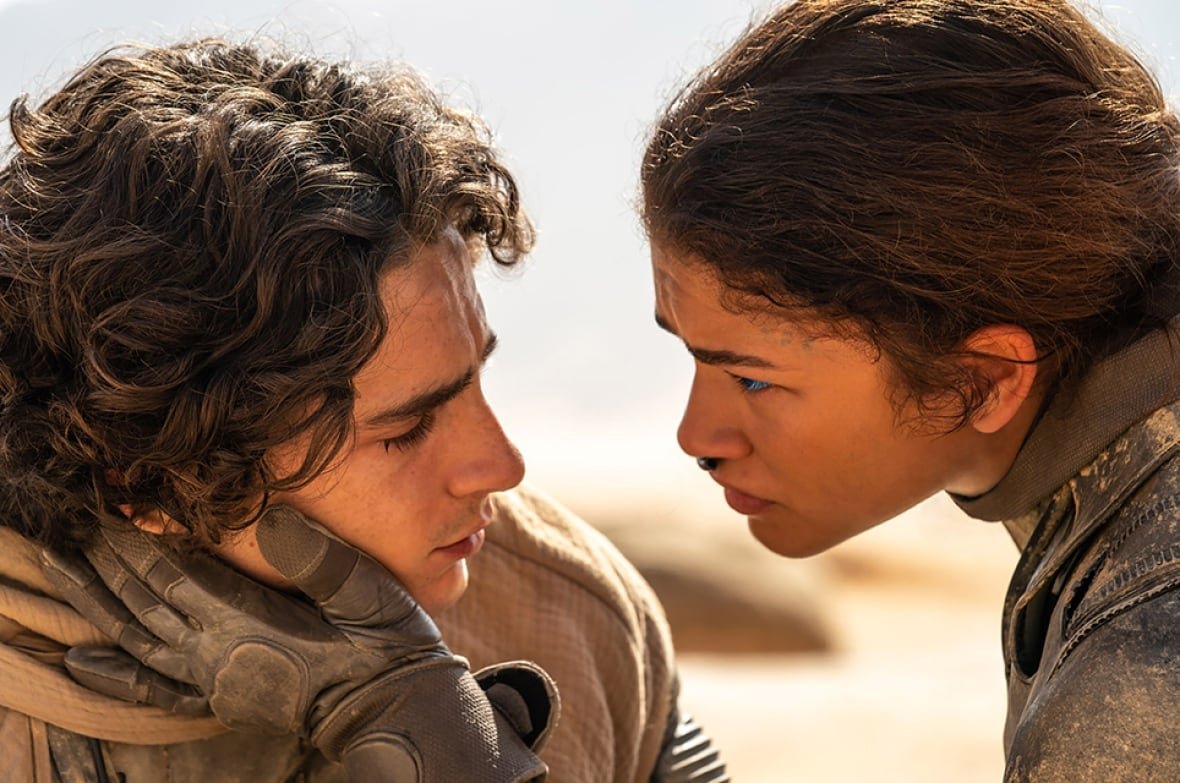 A man and woman stare longingly into each other's eyes. Behind them are desert dunes. 