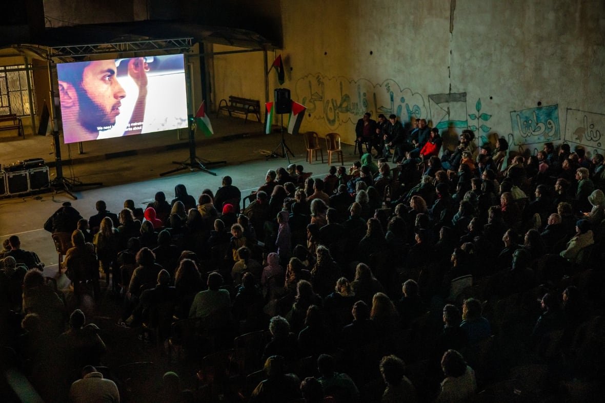 An audience watches a movie projected on a large outdoor screen.