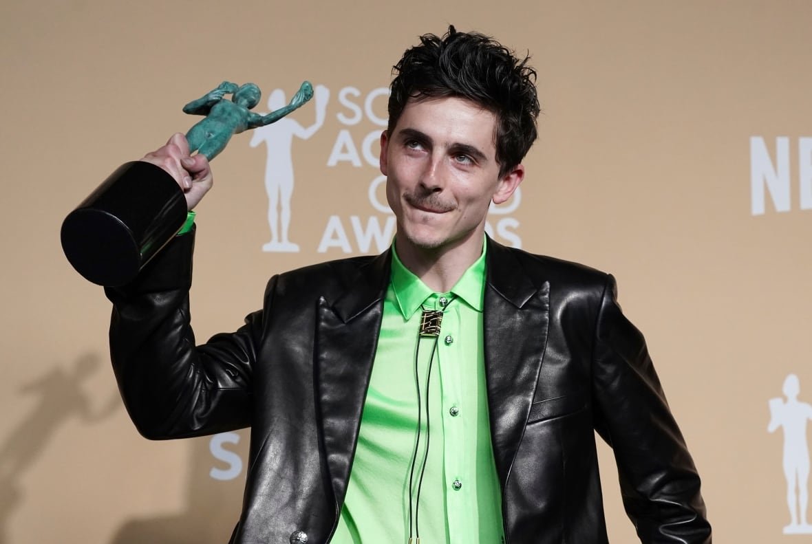 A man wearing a leather blazer and lime green shirt holds a trophy in front of a background that says "Screen Actors Guild Awards"