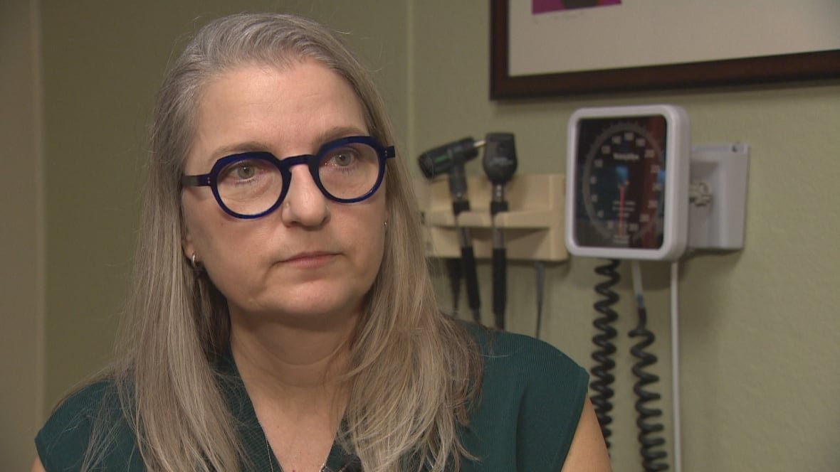 A woman with long, grey hair and dark-framed glasses sits in a room in front of medical equipment mounted on a wall. 