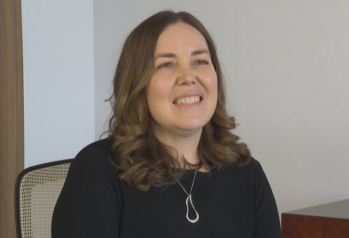 A woman with long, dark hair wearing a black sweater smiles while sitting in an office setting. 