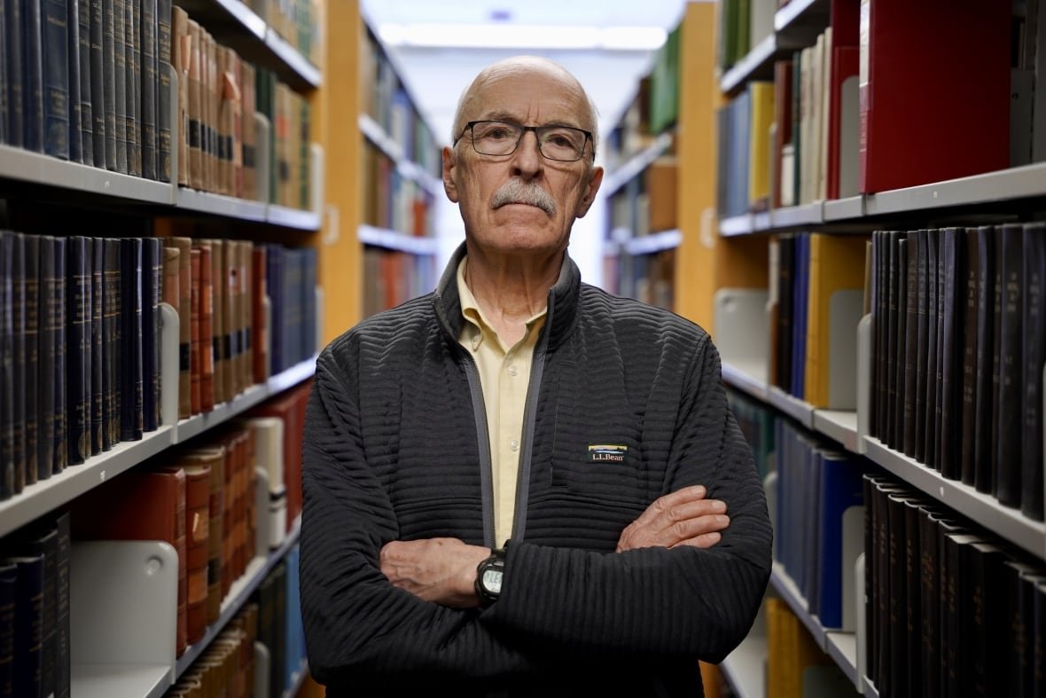 man poses between two rows of books with his arms folded.