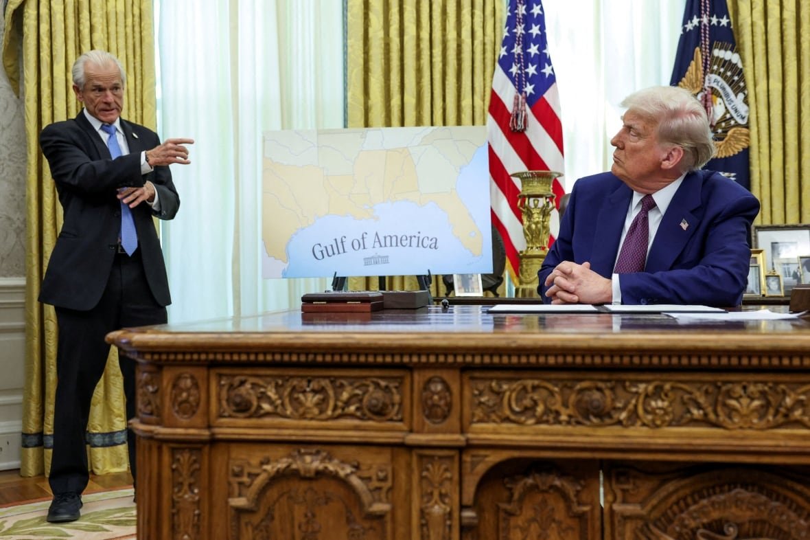 A person standing points to a person sitting at a large desk.