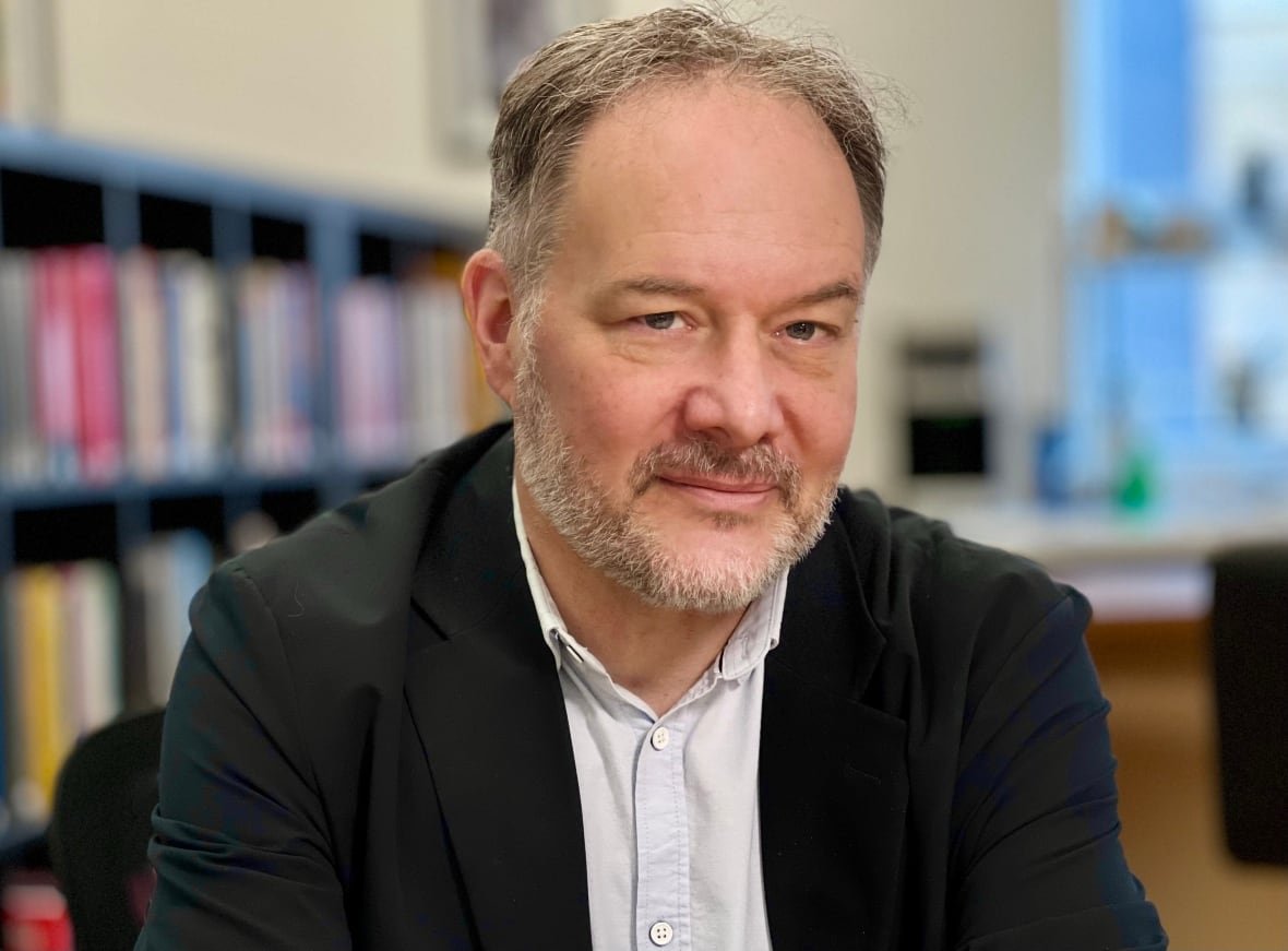 A close-up portrait of a man in a blazer and button-down shirt sitting indoors, with bookshelves and a window seen behind him.