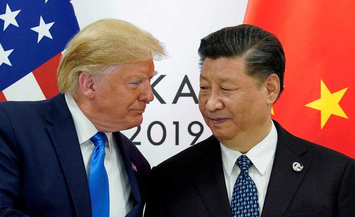 A man with yellow hair and a man with black hair, both wearing dark suits, look at each other.