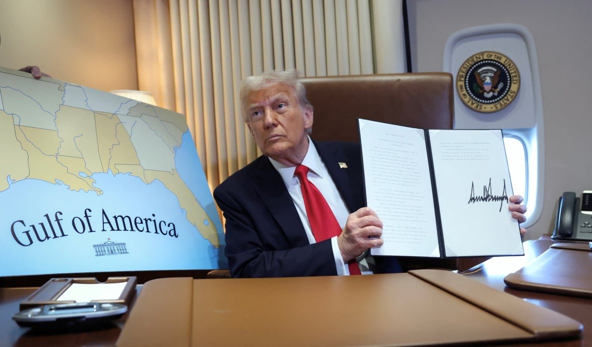 A person sitting at a desk holds a folder displaying a document. To the person's right there is a large map of the southern United States and the words "Gulf of America" over a body of water below the land.