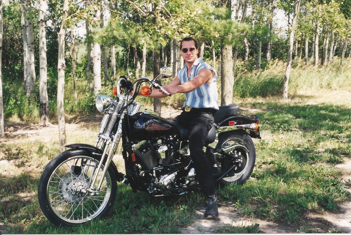 A man in sunglasses and a sleeveless shirt sits on a motorcycle on a sunny summer day.
