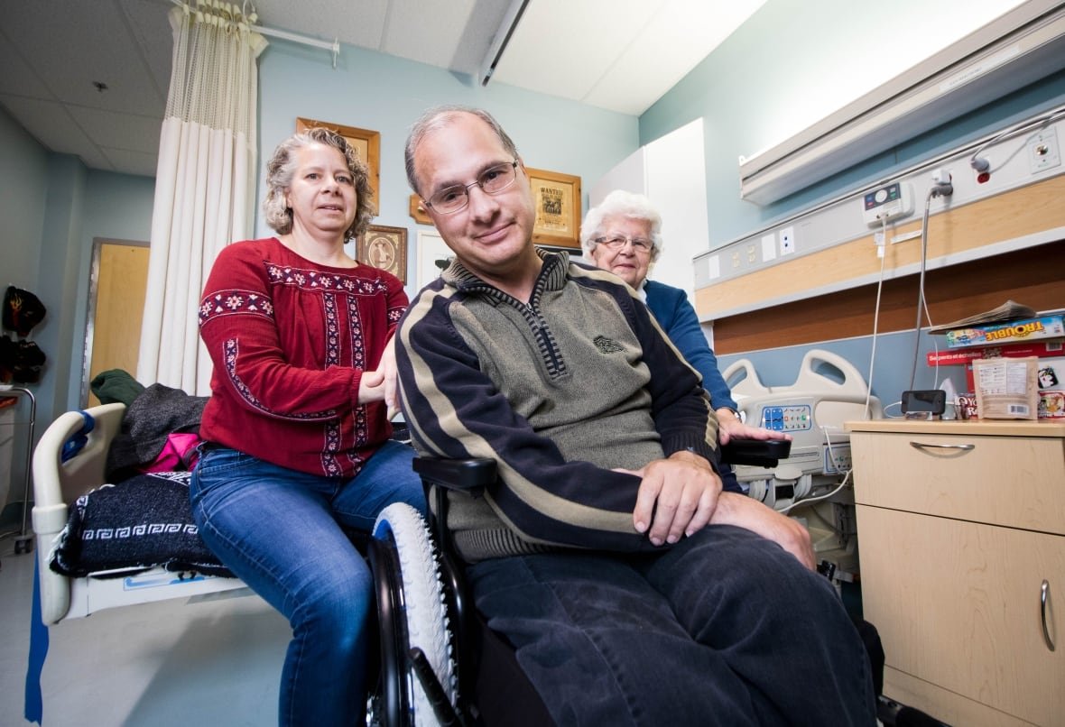 A man sits in a wheelchair with his arms crossed in a hospital room, flanked by two other women.