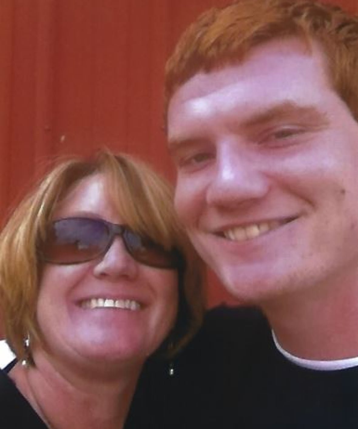In a selfie-style photo, a woman poses with her 20-year-old son, she is wearing sunglasses and both are smiling at the camera.