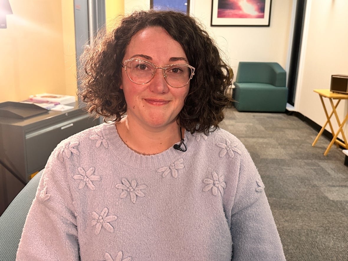 A woman with short curly hair in a blue sweater sits, looking at the camera. 