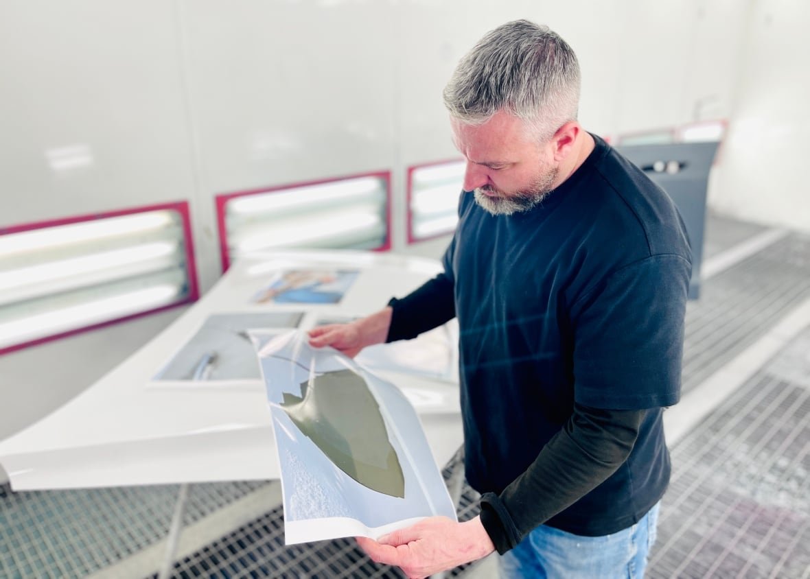 A man in a black T-shirt and jeans looks down, examining the photo of a car with peeling paint.