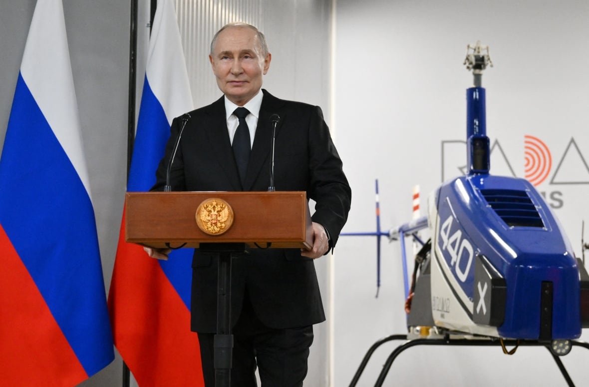 A balding older and cleanshaven Caucasian man wearing a suit and tie stands before a podium. Also shown in the photo are a camera and Russian flags.