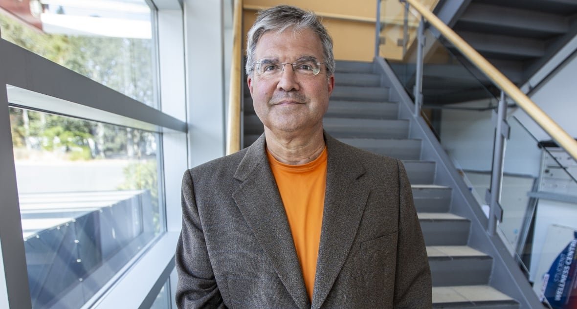 Man with grey hair and glasses wearing a grey blazer over orange shirt standing in front of a flight of stairs in natural light. 