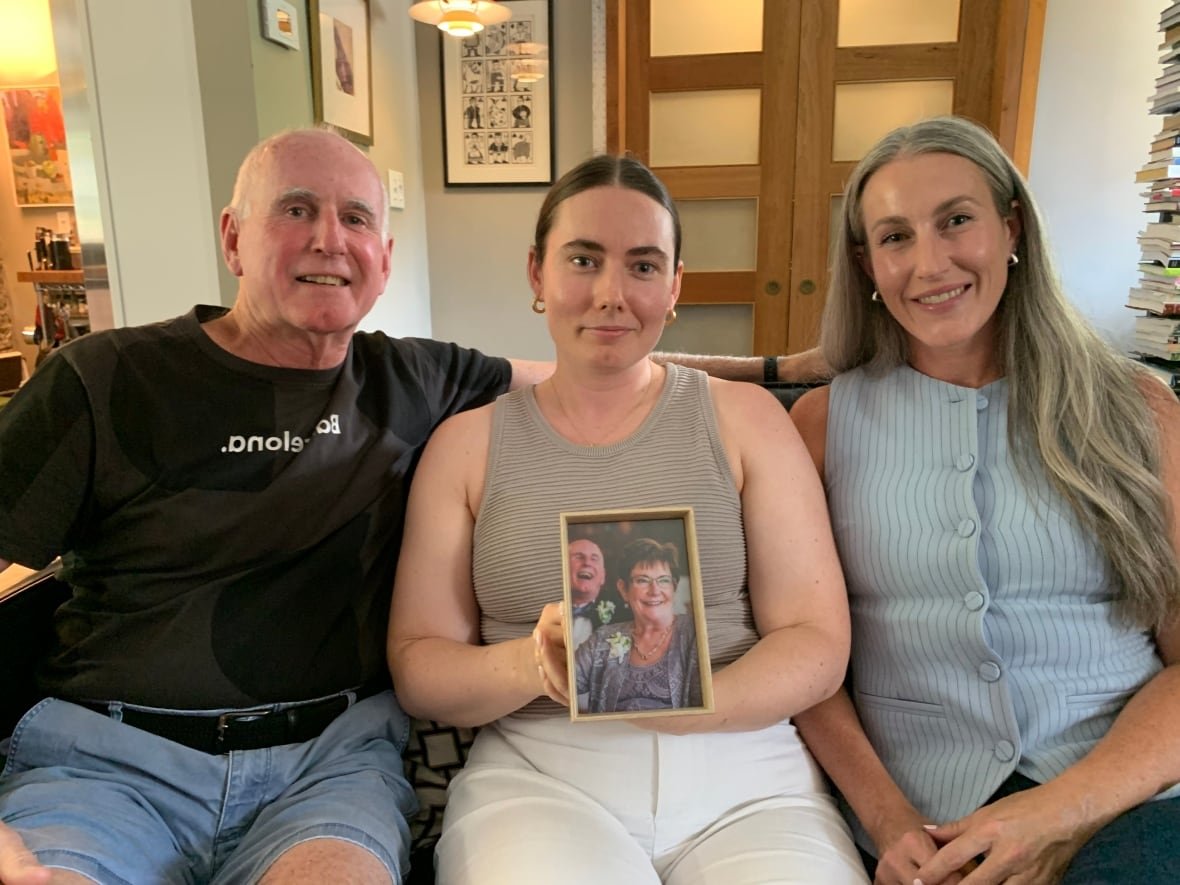 A bald man sits next to two younger women on a sofa. The woman in the middle is holding a portrait of the older man and his wife.