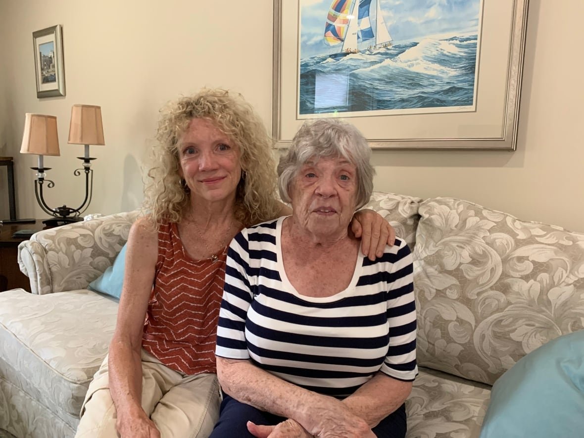 A woman with curly blond hair poses for a portrait with her arm around an older woman next to her on a sofa.