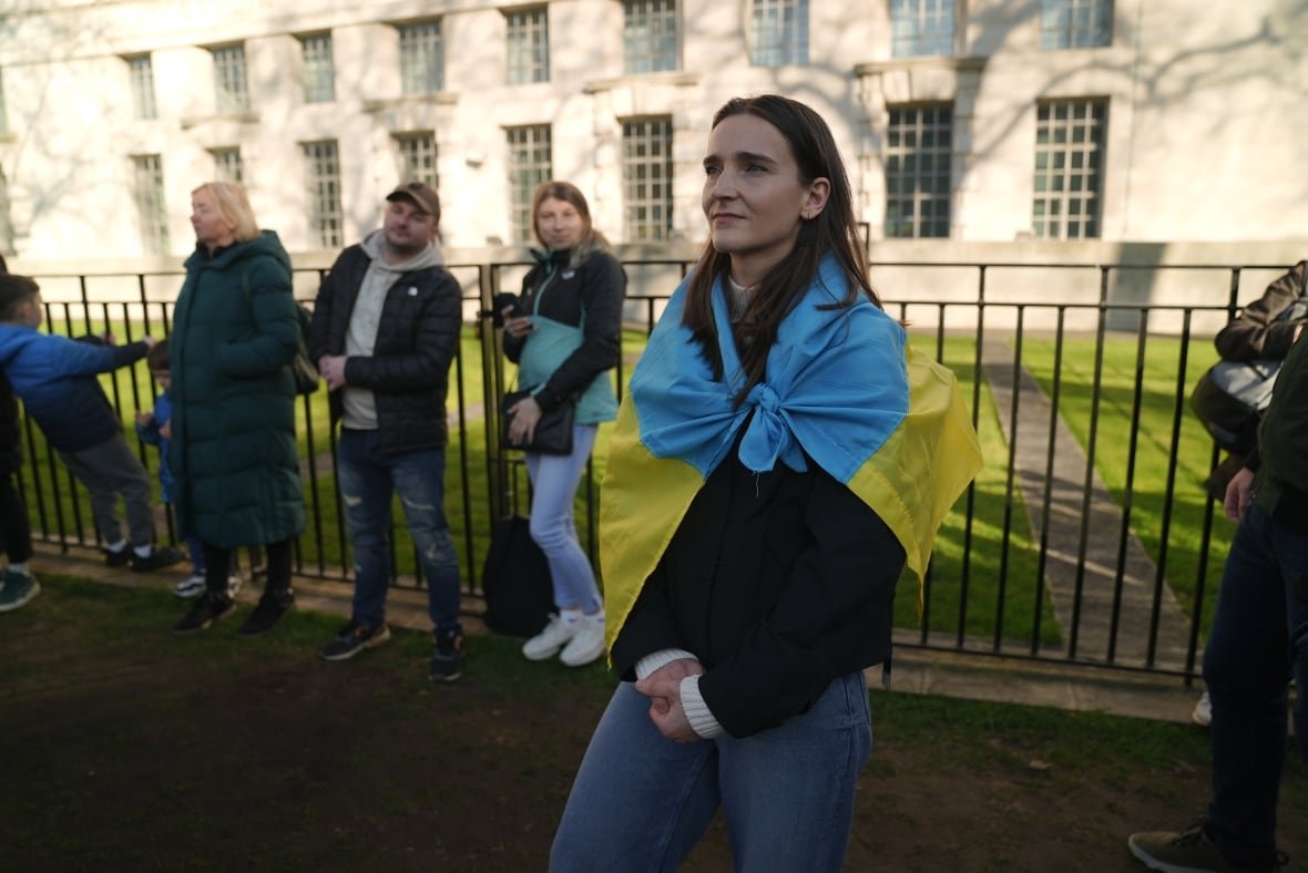 A woman with a flag wrapped around her shoulders watches as people hold a rally.