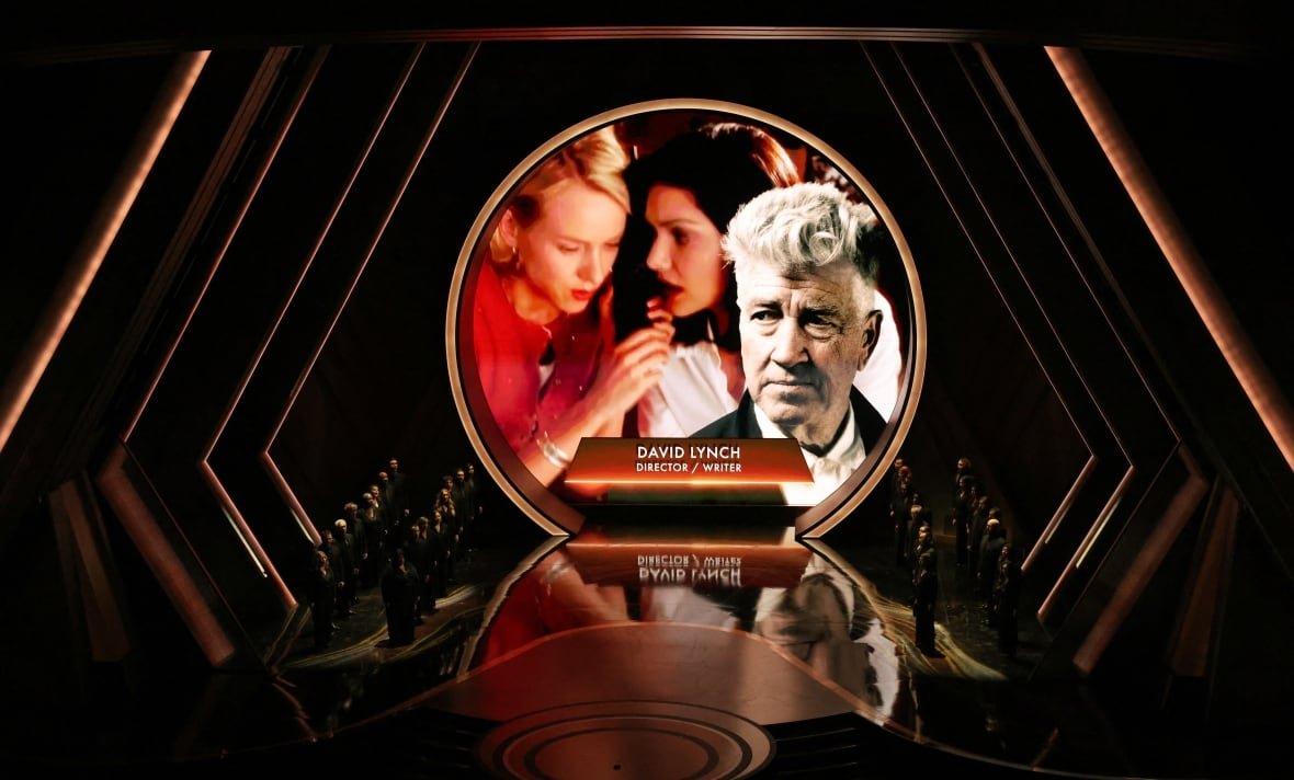 An image of a man with a silver coif of hair is projected on a darkened stage at a theatre. 