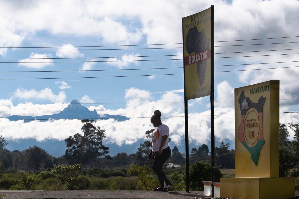A woman walks by signs that  say  Nanyuki. A mount rises in the background