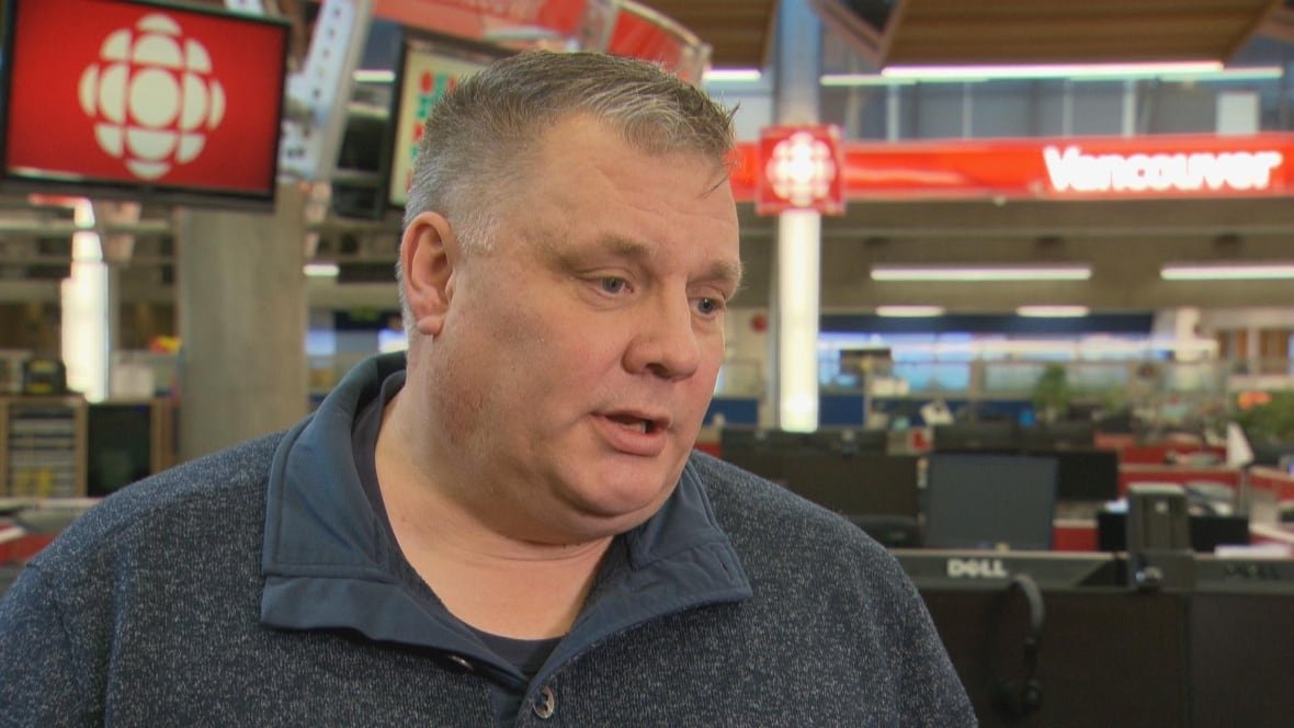 A man wearing a dark blue shirt speaks in a newsroom.