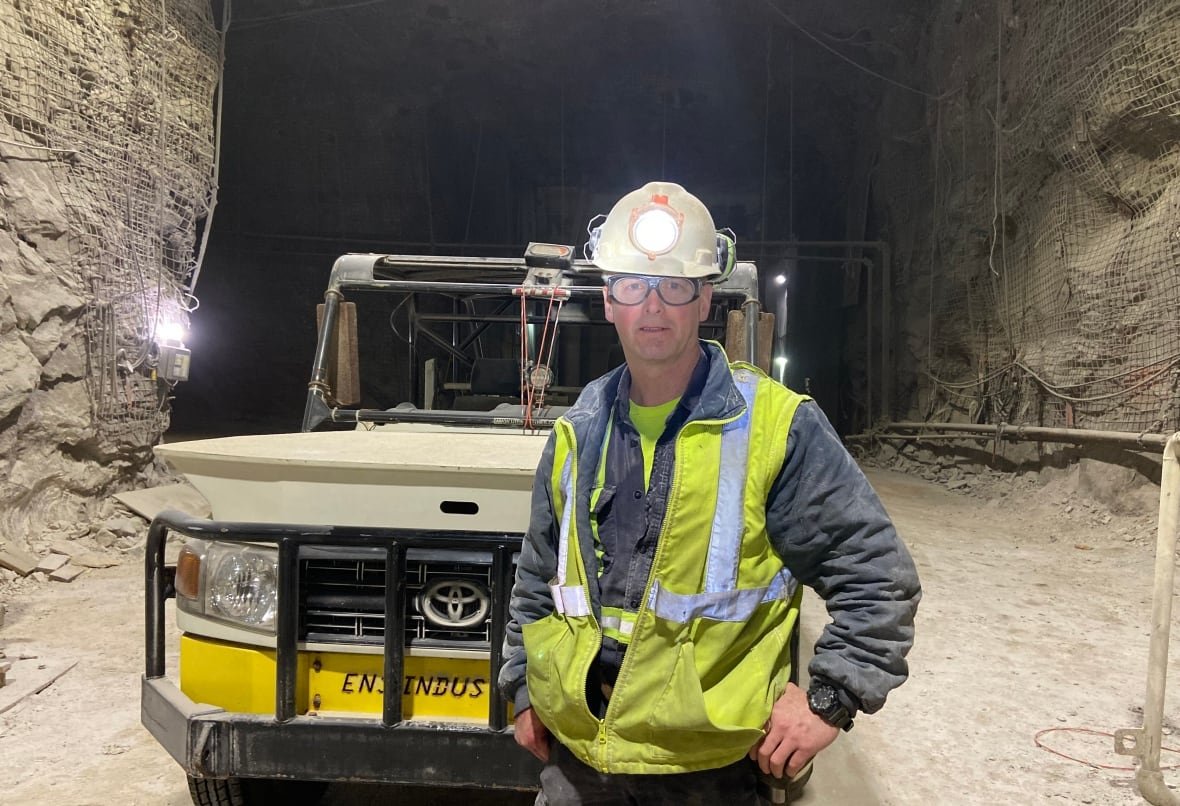 Miner dressed in safety gear wearing hard hat with head lamp is standing in front of Toyota truck inside one of the tunnels in the Picadilly mine.
