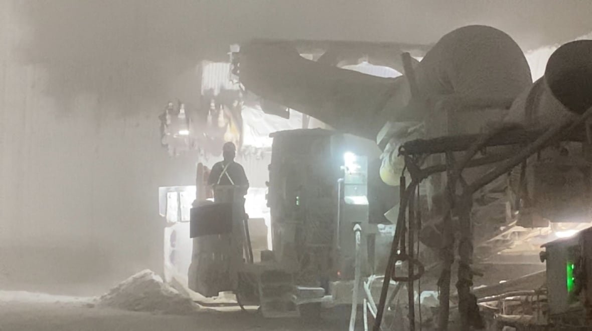 Image of a mining machine with a screw-shaped drill bit grinding away at the wall of the salt deposit cavern. A crew member stands to the left side.
